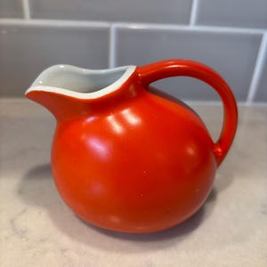 May include: A small, spherical ceramic creamer pitcher with a vibrant orange exterior and a smooth, glossy finish. The pitcher features a curved handle on the side and a white interior lining that extends to the rim of the pouring spout.