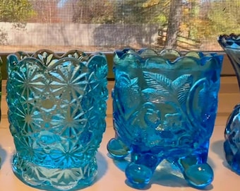 May include: Two vibrant blue glass vessels with distinct textures. The left piece features a repeating starburst pattern with a scalloped rim. The right piece displays a raised fruit and leaf motif, supported by three rounded feet with a scalloped top edge.