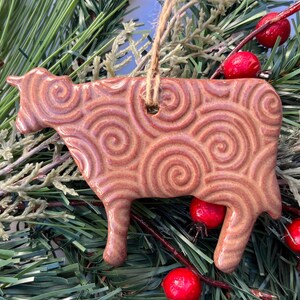 May include: A ceramic cow ornament with a brown glaze and a spiral pattern. The ornament hangs from a twine loop and is surrounded by green pine needles and red berries, creating a festive holiday theme.