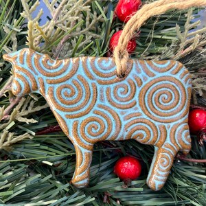 May include: A ceramic cow ornament with a light blue base and brown spiral design. The ornament is hanging from a natural twine string. The cow is surrounded by green pine needles and red berries, creating a festive holiday theme.