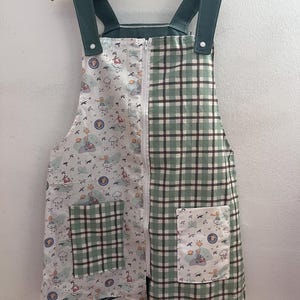 Peut inclure: Une robe salopette à carreaux verts et blancs avec une fermeture éclair blanche et des bretelles vertes. Le côté gauche de la robe présente un imprimé fantaisiste avec des nuages, des soleils et des personnages. La robe a deux poches avant.