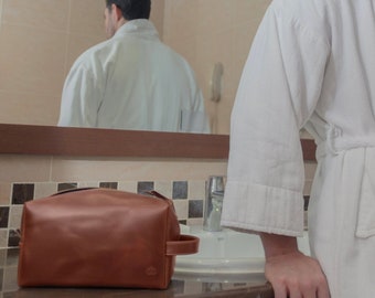 May include: A brown leather toiletry bag with a zipper closure and a handle. The bag is sitting on a counter in a bathroom.