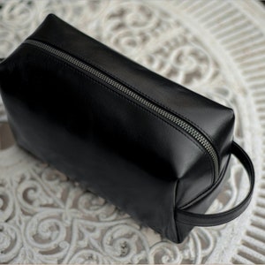 May include: Black rectangular travel bag with a silver zipper and a small handle. The bag is made of a smooth, shiny material and sits on a white, ornate table.