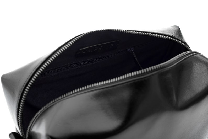 May include: Black leather cosmetic bag with a silver zipper. The bag is open, revealing a dark interior with a small zippered pocket. The brand name is visible inside the bag.