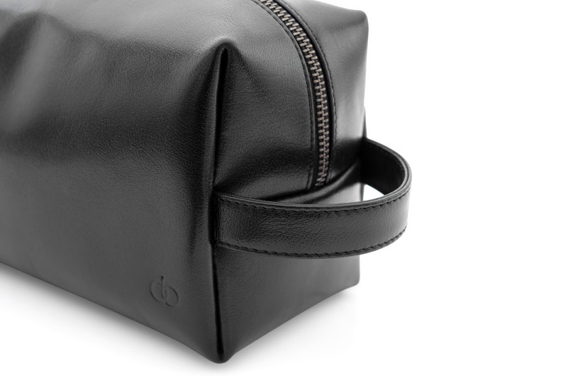 May include: A black leather toiletry bag with a silver zipper and a handle. The bag is square-shaped and features a small embossed logo on the front. The bag is designed for travel and personal care items.