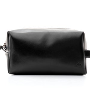May include: A black leather toiletry bag with a zipper closure and a small handle on one side. The bag has a rectangular shape and a smooth, glossy finish. A small logo is visible on the front.