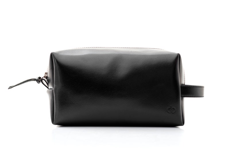 May include: A black leather toiletry bag with a zipper closure and a small handle on one side. The bag has a rectangular shape and a smooth, glossy finish. A small logo is visible on the front.