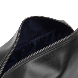 May include: Black leather toiletry bag with a silver zipper. The interior is lined with dark blue fabric and features a small pocket. The bag is open, revealing the inside.