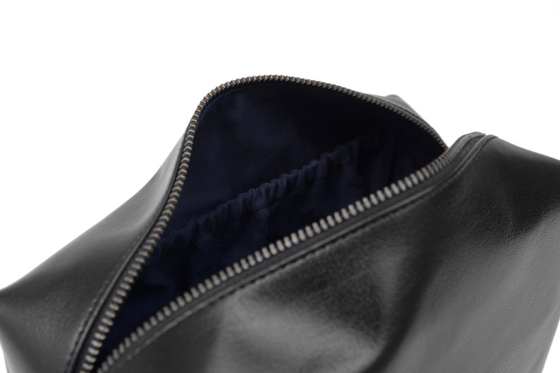 May include: Black leather toiletry bag with a silver zipper. The interior is lined with dark blue fabric and features a small pocket. The bag is open, revealing the inside.