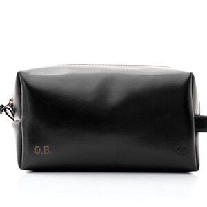 May include: Black leather toiletry bag with a rectangular shape, a zipper closure, and a small handle on one side. The bag features the initials "O.B." in the lower left corner and a logo on the lower right corner. The bag is designed for travel and personal care items.
