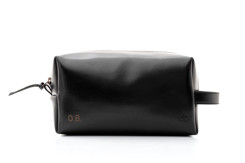 May include: Black leather toiletry bag with a rectangular shape, a zipper closure, and a small handle on one side. The bag features the initials "O.B." in the lower left corner and a logo on the lower right corner. The bag is designed for travel and personal care items.