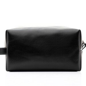 May include: A black leather toiletry bag with a rectangular shape. The bag has a zipper closure, a small handle on one side, and a leather pull tab on the zipper. The bag is designed for travel and personal care items.