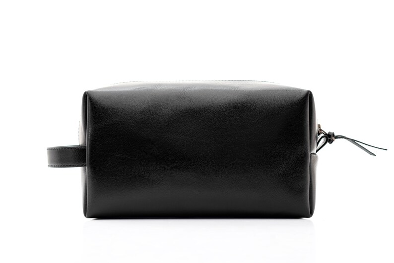 May include: A black leather toiletry bag with a rectangular shape. The bag has a zipper closure, a small handle on one side, and a leather pull tab on the zipper. The bag is designed for travel and personal care items.