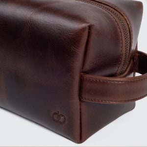 May include: A brown leather travel wash bag with a zip fastening and a leather handle. The bag has a small embossed logo on the front.