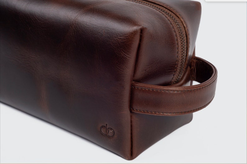 May include: A brown leather travel wash bag with a zip fastening and a leather handle. The bag has a small embossed logo on the front.
