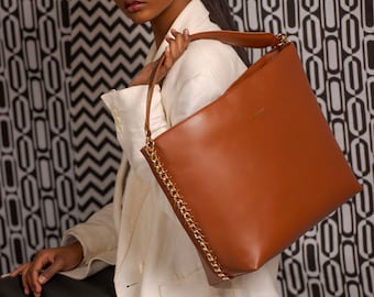 May include: A woman wearing a white blazer and black pants holds a brown leather tote bag with a gold chain strap. The bag has a gold logo on the front.