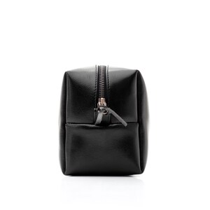 May include: A black, rectangular cosmetic bag with a zipper closure. The bag is made of a smooth, leather-like material and has a small pocket on the front. The zipper pull is a small leather tab.