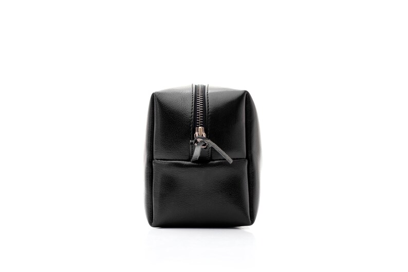 May include: A black, rectangular cosmetic bag with a zipper closure. The bag is made of a smooth, leather-like material and has a small pocket on the front. The zipper pull is a small leather tab.