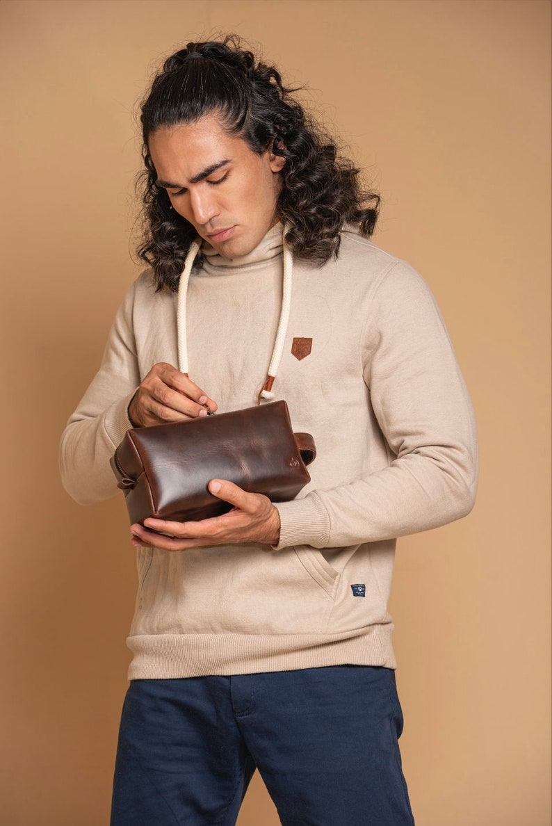 May include: A person holding a brown leather wash bag with a white rope handle. The bag is closed and the person is looking down at it.