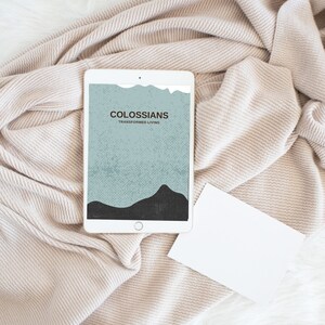 May include: A white tablet displaying the text "COLOSSIANS TRANSFORMED LIVING" over a blue and black mountain graphic. The tablet rests on a beige knitted blanket, with a blank white card beside it.