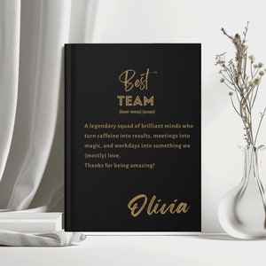 May include: Black notebook with gold lettering that reads "Best Team [best-teem] (noun) A legendary squad of brilliant minds who turn caffeine into results, meetings into magic, and workdays into something we (mostly) love. Thanks for being amazing! Olivia"