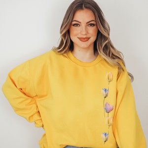 Peut inclure: Un sweat-shirt jaune avec une rang&eacute;e verticale de tulipes de style aquarelle dans des tons de rose, bleu et jaune sur le c&ocirc;t&eacute; droit.