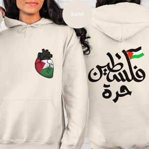 Może przedstawiać: Szarobrązowa bluza z kapturem z sercem z flagą palestyńską z przodu i arabskim tekstem "فلسطين حرة" z flagą palestyńską z tyłu.
