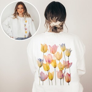 Peut inclure: Sweat-shirt blanc avec un motif aquarelle de tulipes rose pastel, jaunes et bleues au dos. Le devant du sweat-shirt est blanc uni.