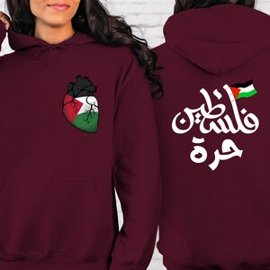 Może przedstawiać: Bordowa bluza z kapturem z nadrukiem w kształcie serca z flagą palestyńską z przodu i arabskim napisem "Palestyna" z flagą palestyńską z tyłu.