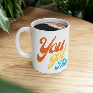 May include: White ceramic coffee mug with a black handle. The mug has the text "You got this" written in a colorful, retro font. The text is in orange, yellow, and blue.