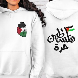 Może przedstawiać: Biała bluza z kapturem z sercem w kształcie palestyńskiej flagi z przodu i arabskim napisem "Palestyna" z palestyńską flagą z tyłu.