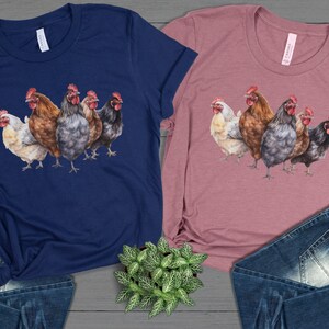 Puede incluir: Dos camisetas, una azul marino y otra malva, cada una con una ilustración en acuarela de cinco gallinas. Las gallinas son de varios colores, incluyendo blanco, marrón, gris y negro, con crestas rojas. Las camisetas se muestran con vaqueros azules y una pequeña planta.