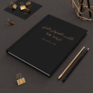 以下が含まれることがあります： 「Aktib al-afwan al-ladhi  teredeh  hna」と「Aktib ma teredh hna」という金色の文字が書かれた黒いノート。ノートは濃いグレーの表面に置かれ、金色の鉛筆と黒い鉛筆が2本、金色のクリップが1つあります。
