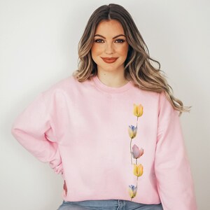 Peut inclure: Un sweat-shirt rose avec une ligne verticale de tulipes aquarelle dans des tons de jaune, bleu et violet.