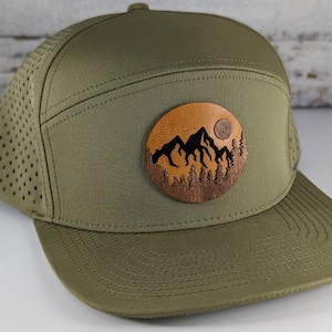 May include: A green mesh and fabric baseball cap with a brown leather patch featuring a mountain silhouette and a full moon.