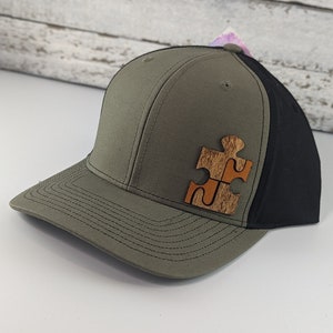 May include: A baseball cap with a green front panel and a black mesh back. The cap features a wooden puzzle piece design on the front. The hat is sitting on a white surface.