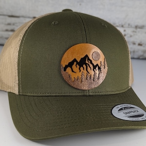 May include: A green and tan trucker hat with a brown leather patch featuring a silhouette of mountains and a full moon. The patch has a tan background.