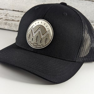 May include: Black trucker hat with a wood-burned patch featuring a mountain range and trees. The patch is in a circle with a white background and a sun.