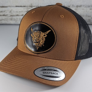 May include: A brown and black trucker hat with a black leather patch featuring a gold embroidered Highland cow head. The hat has a black mesh back and a brown front with a black brim. The hat has a sticker on the bottom that says "The Authentic Snapback Yupoong Since 1974."