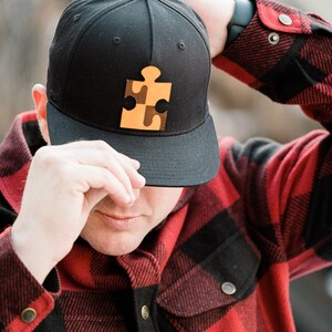 May include: A person wearing a black and red plaid shirt with a black baseball cap. The cap has a yellow puzzle piece design on the front.