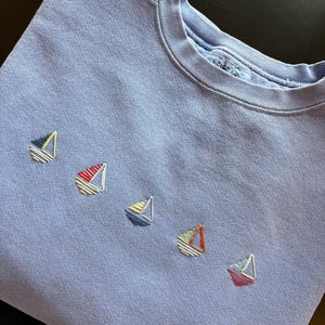 Op de afbeelding: Een periwinkle blauwe sweatshirt met vijf geborduurde zeilbootontwerpen in verschillende kleuren. De zeilboten hebben witte, rode, gele en oranje accenten. De crew neck sweatshirt is gemaakt van een zacht materiaal.