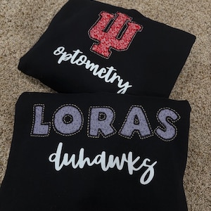 Puede incluir: Dos sudaderas negras. La sudadera superior tiene un diseño rojo y blanco con la palabra "optometry" en escritura blanca. La sudadera inferior tiene la palabra "LORAS" en letras grises y "duhawks" en escritura blanca.
