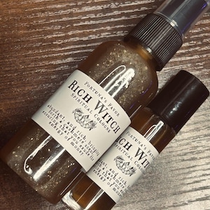 Puede incluir: Dos botellas de "Rich Witch Spiritual Cologne" con etiquetas blancas y texto negro. Las botellas están llenas de un líquido ámbar brillante. Las etiquetas incluyen texto sobre el aroma y la energía del dinero. Sobre una superficie de madera.