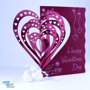 May include: A burgundy card with a 3D pop-up heart design. The heart is made of multiple layers of paper, each with a different size and shape. The card is decorated with white hearts and the text "Happy Valentines Day".