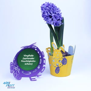 Op de afbeelding: Een paarse en gele papieren plantenbak met een konijnenontwerp en een hyacintenplant. De plantenbak heeft een vinylfolie voor optionele vochtbescherming. De tekst "Vinylfolie (optionaler Feuchtigkeits-schutz)" staat op de plantenbak gedrukt.
