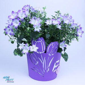 Op de afbeelding: Een paarse plantenpot met een tulpenontwerp, gevuld met een bloeiende plant met lichtpaarse bloemen. De pot heeft een klein roze label met een lieveheersbeestje-ontwerp.