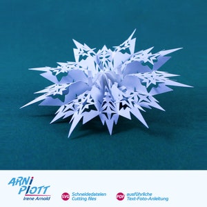 May include: A white paper snowflake with a detailed, intricate design. The snowflake is folded and has a three-dimensional appearance. The snowflake is on a teal background.