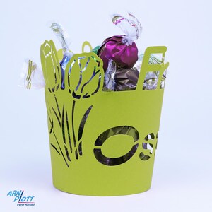 Op de afbeelding: Een groene papieren paaskorft met een tulpenontwerp en het woord "Pasen" uitgesneden. De korf is gevuld met verschillende chocolaatjes.