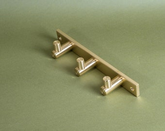 May include: A gold metal wall-mounted hook rack with three hooks. The rack is mounted on a green background.