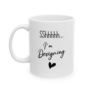 May include: White ceramic mug with a curved handle. The mug features the text "SShhhhh... I'm Designing" in black script, along with a black heart graphic. The mug is designed for designers and creatives.